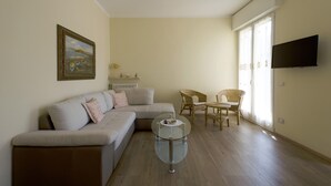 Een woonkamer met een bank, een glazen salontafel, rieten stoelen, een schilderij en een aan de muur bevestigde televisie.