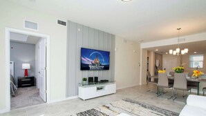 Una sala de estar moderna con un televisor de pantalla plana montado en la pared, una zona de comedor con mesa y sillas, y un pasillo que conduce a un dormitorio.