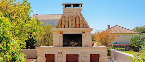 A stone-built outdoor kitchen with a chimney, surrounded by greenery and a tiled roofed building in the background.