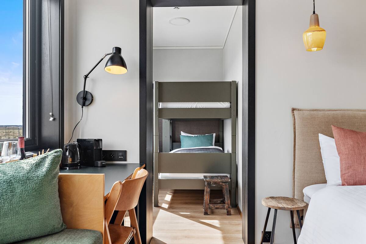 Une chambre d’hôtel moderne équipée d’un lit superposé, d’un bureau avec une chaise, d’un petit tabouret et d’une table de chevet avec une lampe.