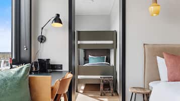 A modern hotel room with a bunk bed, a desk with a chair, a small stool, and a bedside table with a lamp.