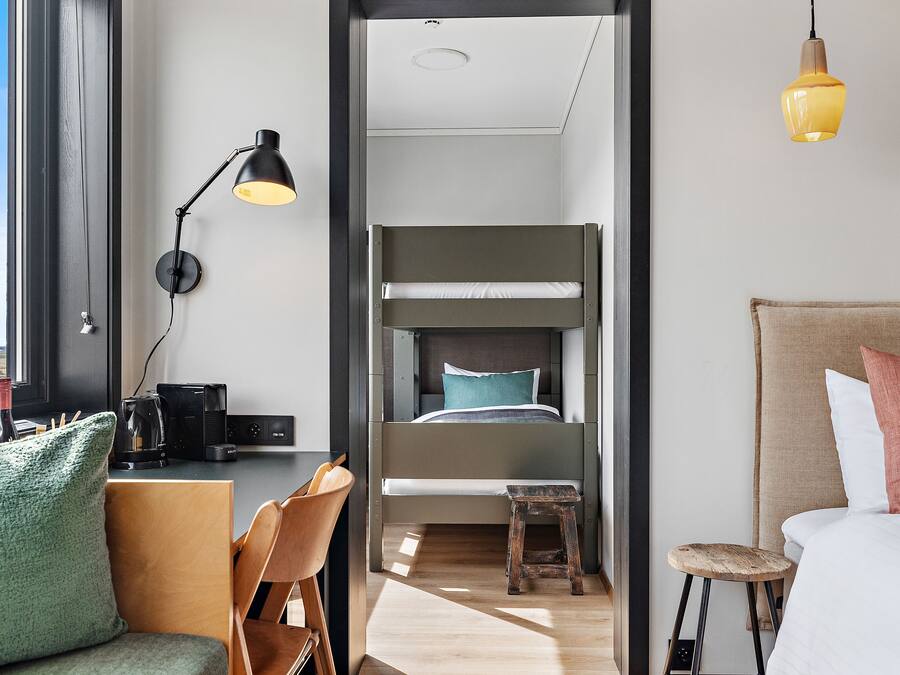 A modern hotel room with a bunk bed, a desk with a chair, a small stool, and a bedside table with a lamp.