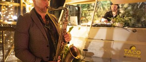 Uma pessoa tocando saxofone em frente a um food truck com uma placa que diz 'TACO VAGINN'.