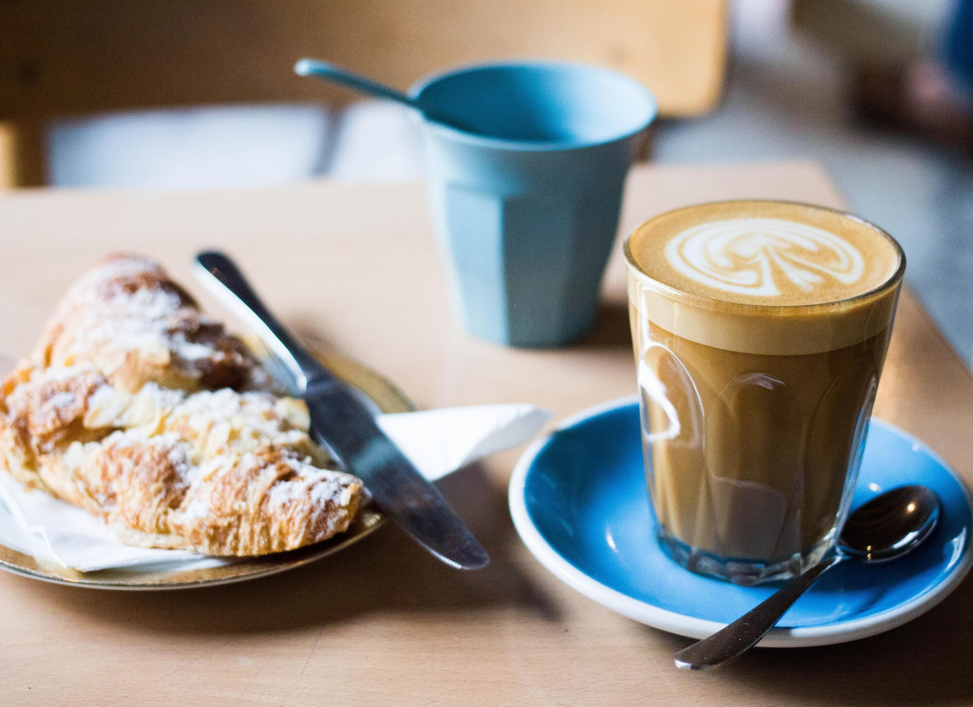 A table with a pastry, a cup of coffee, a spoon, and a blue cup.