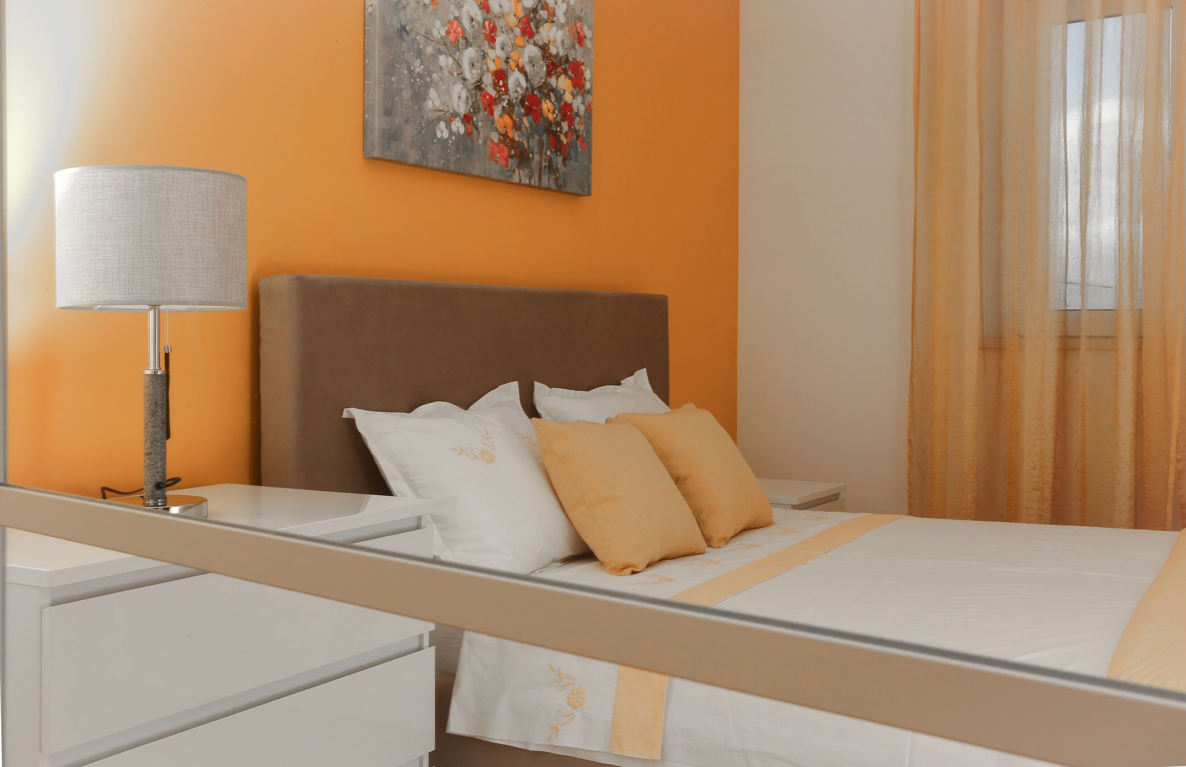 A neatly made bed with white and orange bedding, a bedside lamp, and a painting on the wall.