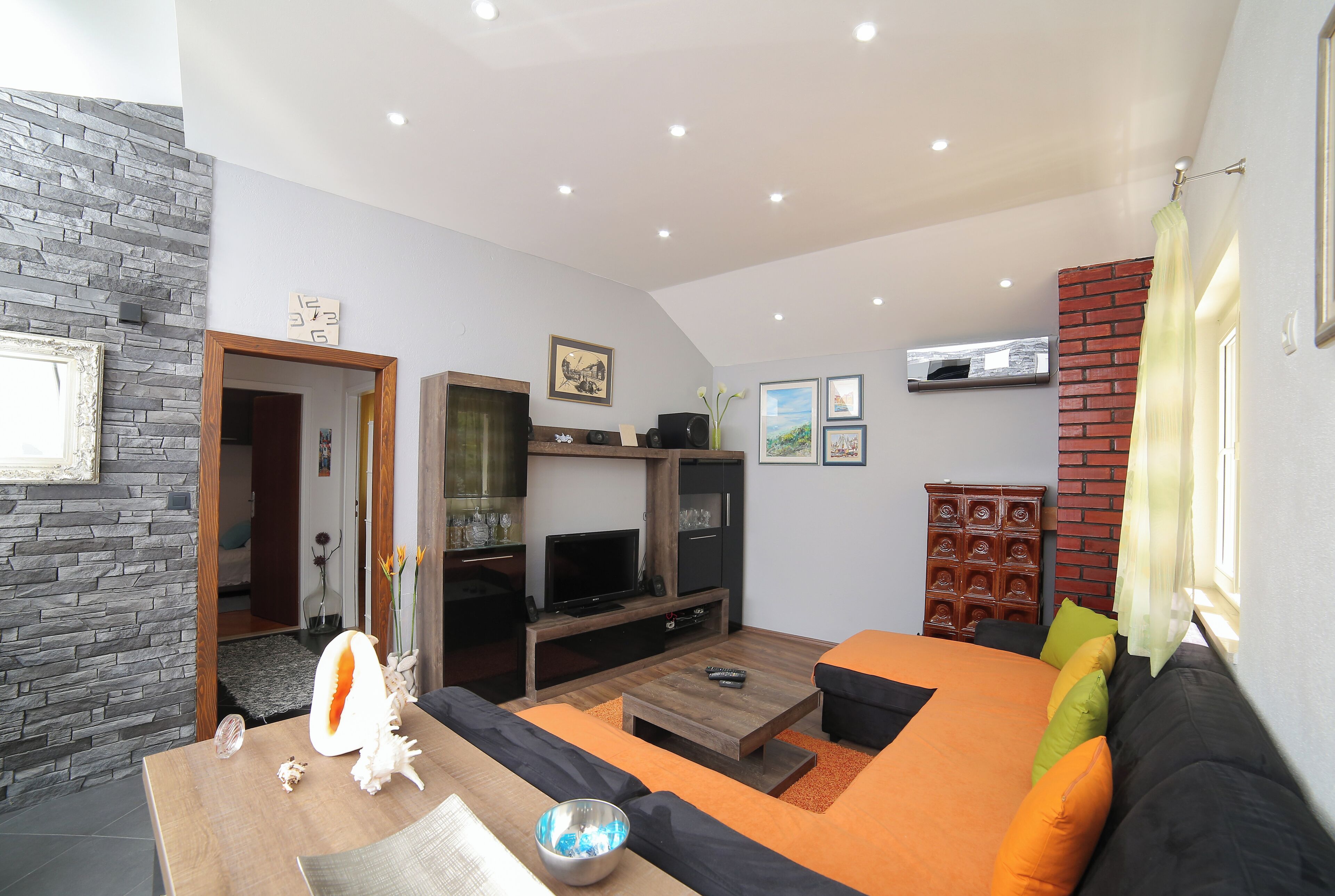 A modern living room with a stone wall, a wooden coffee table, a sofa with orange cushions, a TV on a wooden stand, and a brick accent wall.