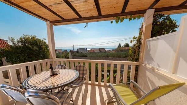 A covered outdoor patio with a round table and chairs, a yellow folding chair, and a view of houses and greenery in the background. - Apartments Magdalena (Bol)