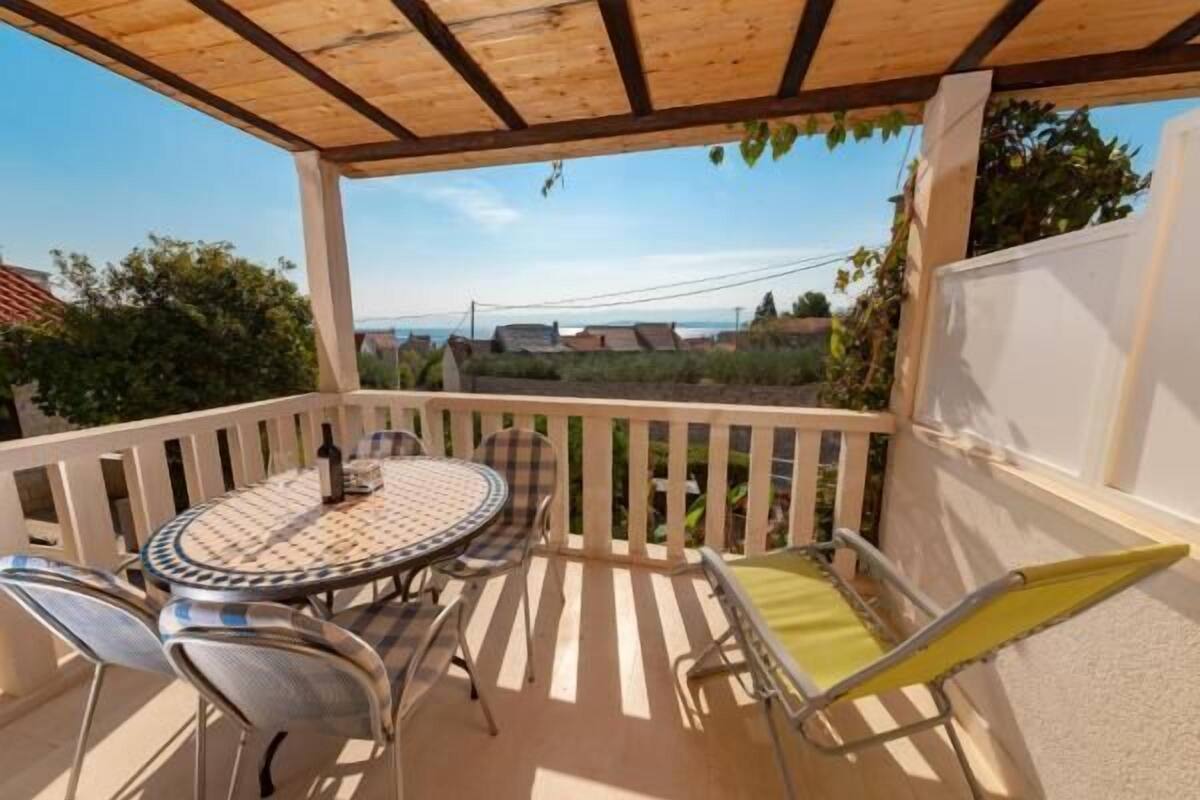 Um alpendre coberto com uma mesa redonda e cadeiras, uma cadeira dobrável amarela, e vista para casas e vegetação ao fundo.