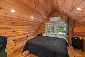 Un dormitorio acogedor con techo inclinado, paredes de madera, una cama con colcha oscura, una mesita de noche con lámpara y una ventana con vistas a árboles.