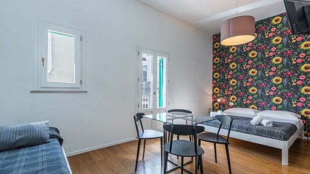 Una habitación con cama, una mesa de comedor con sillas, una ventana y un papel tapiz floral en la pared.