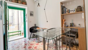 A compact living space with a glass dining table, a black chair, a bookshelf, and a small balcony with a green door. - Locanda LoSteri (Ragusa)