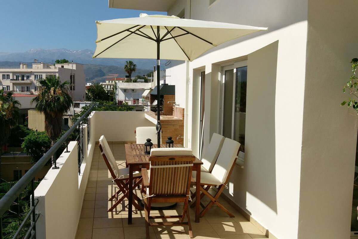 A balcony with a table and chairs, a large umbrella, and a view of the city and mountains in the background.
