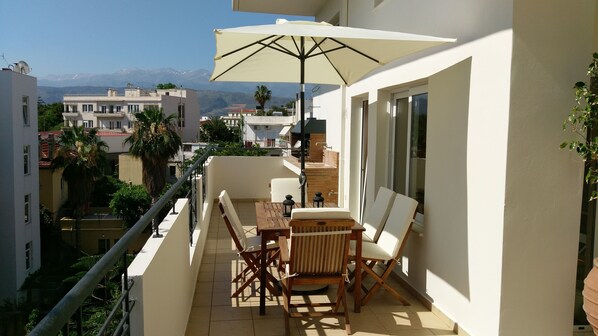 A balcony with a table and chairs, a large umbrella, and a view of the city and mountains in the background.