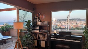 A living room with a piano, a bookshelf with vinyl records, a lamp, and a view of a cityscape through large windows. - ChaniaStay Central Apartments (Chania)