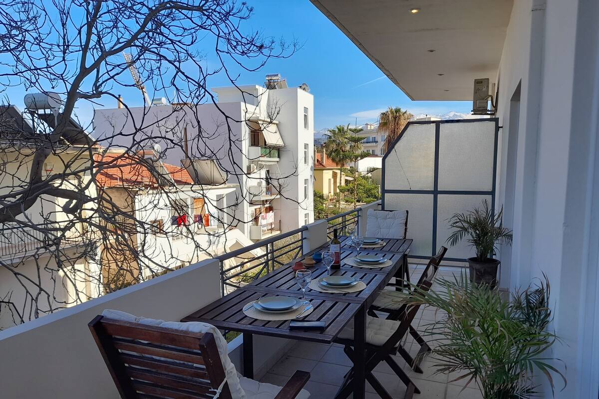 A balcony with a dining table set for two, surrounded by wooden chairs and potted plants.
