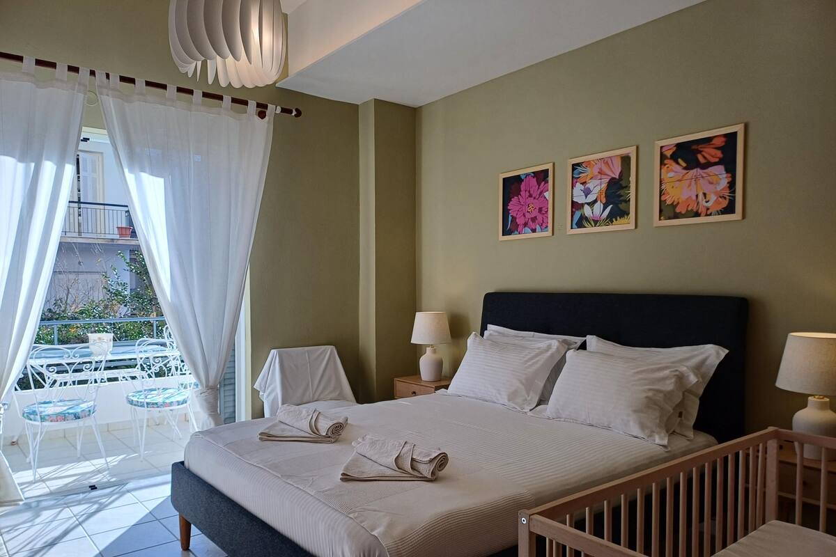 A bedroom with a bed, two bedside tables with lamps, a crib, a balcony with a table and chairs, and three floral paintings on the wall.
