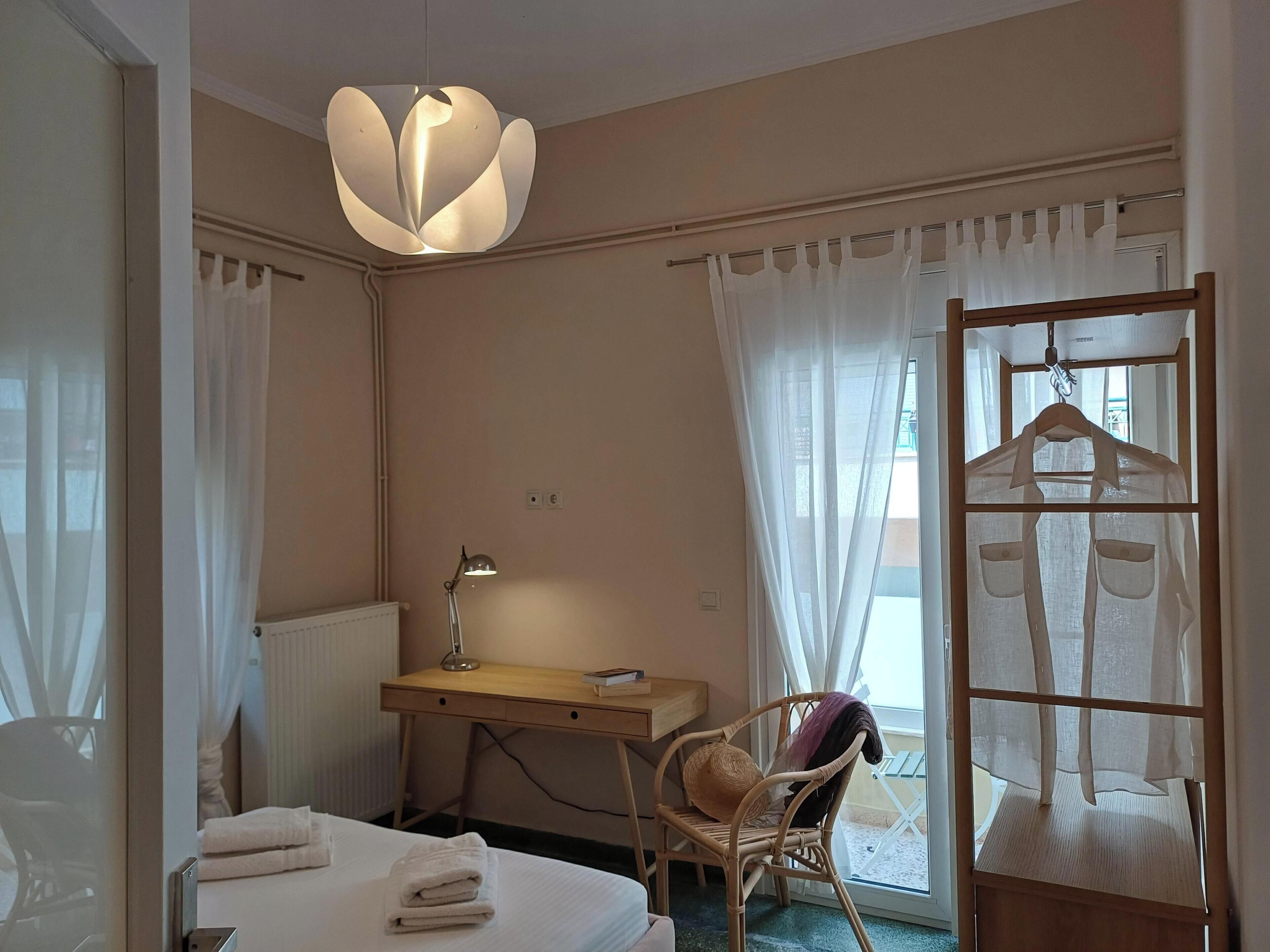 Een hotelkamer met een bed, een bureau, een stoel, een kledingroede en een raam met dunne gordijnen.