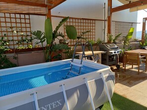 An outdoor swimming pool with a ladder, surrounded by plants and a barbecue area.