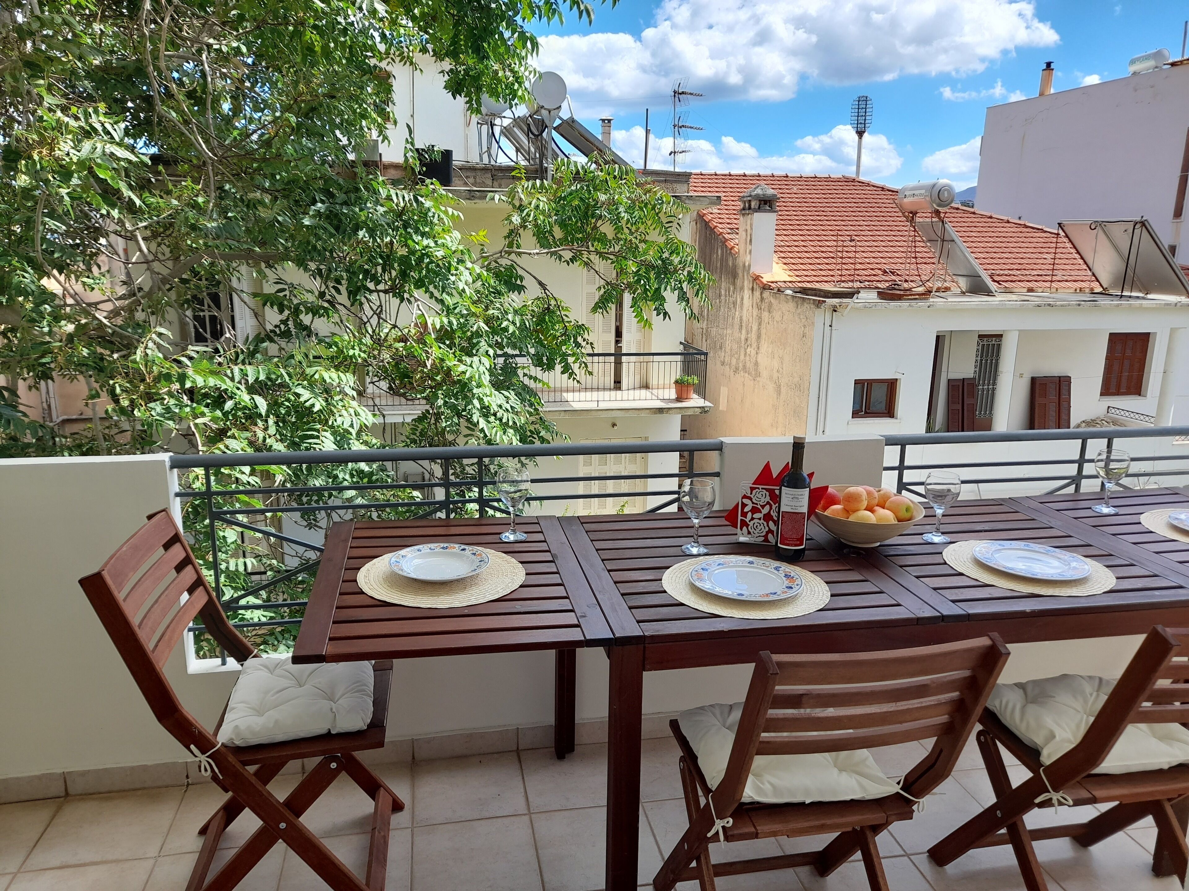 A wooden table set for four with plates, a wine bottle, and a bowl of fruit on a balcony overlooking a cityscape.
