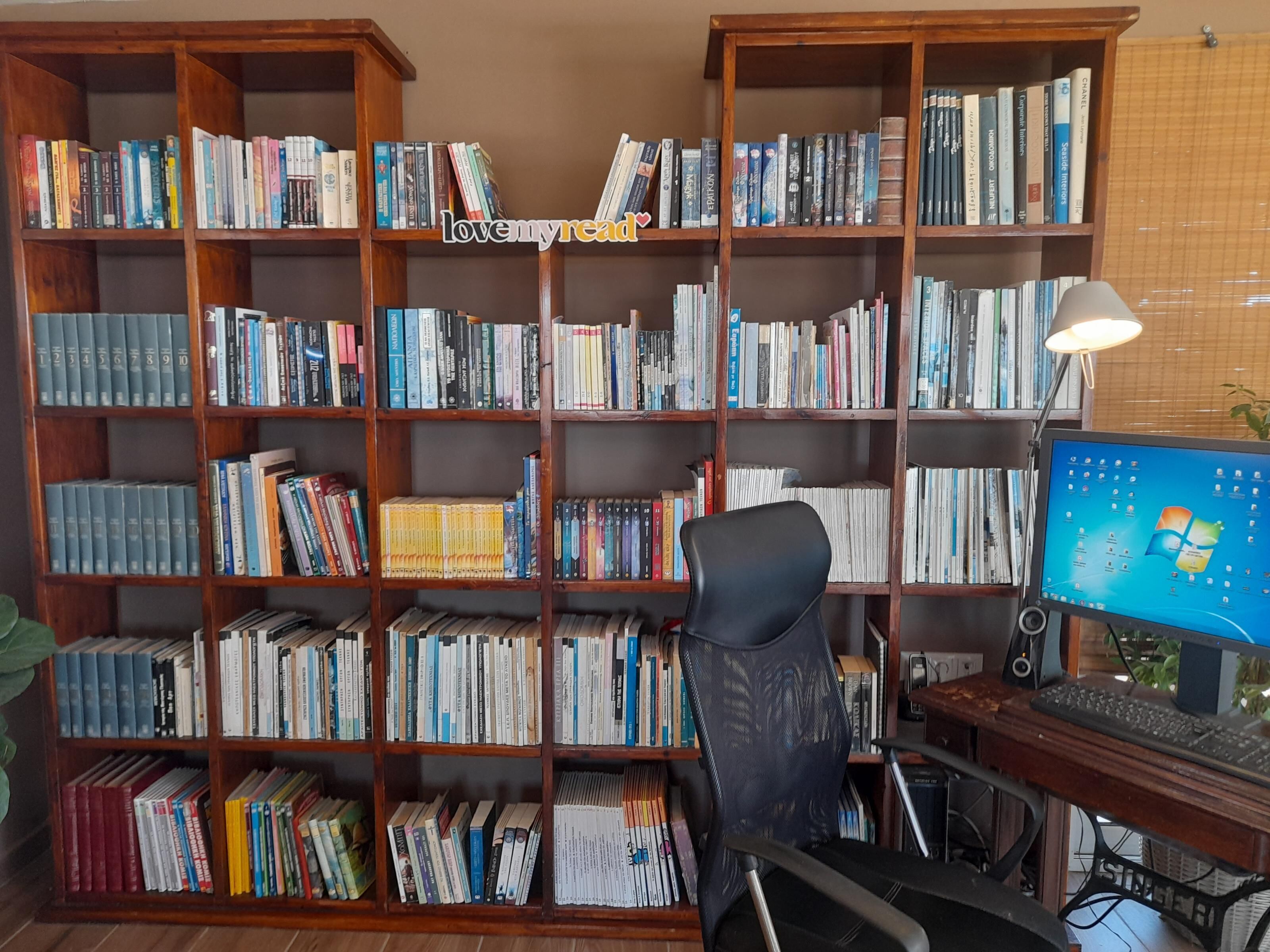 A home office with a wooden bookshelf filled with books, a black office chair, a desk with a computer monitor, and a lamp.