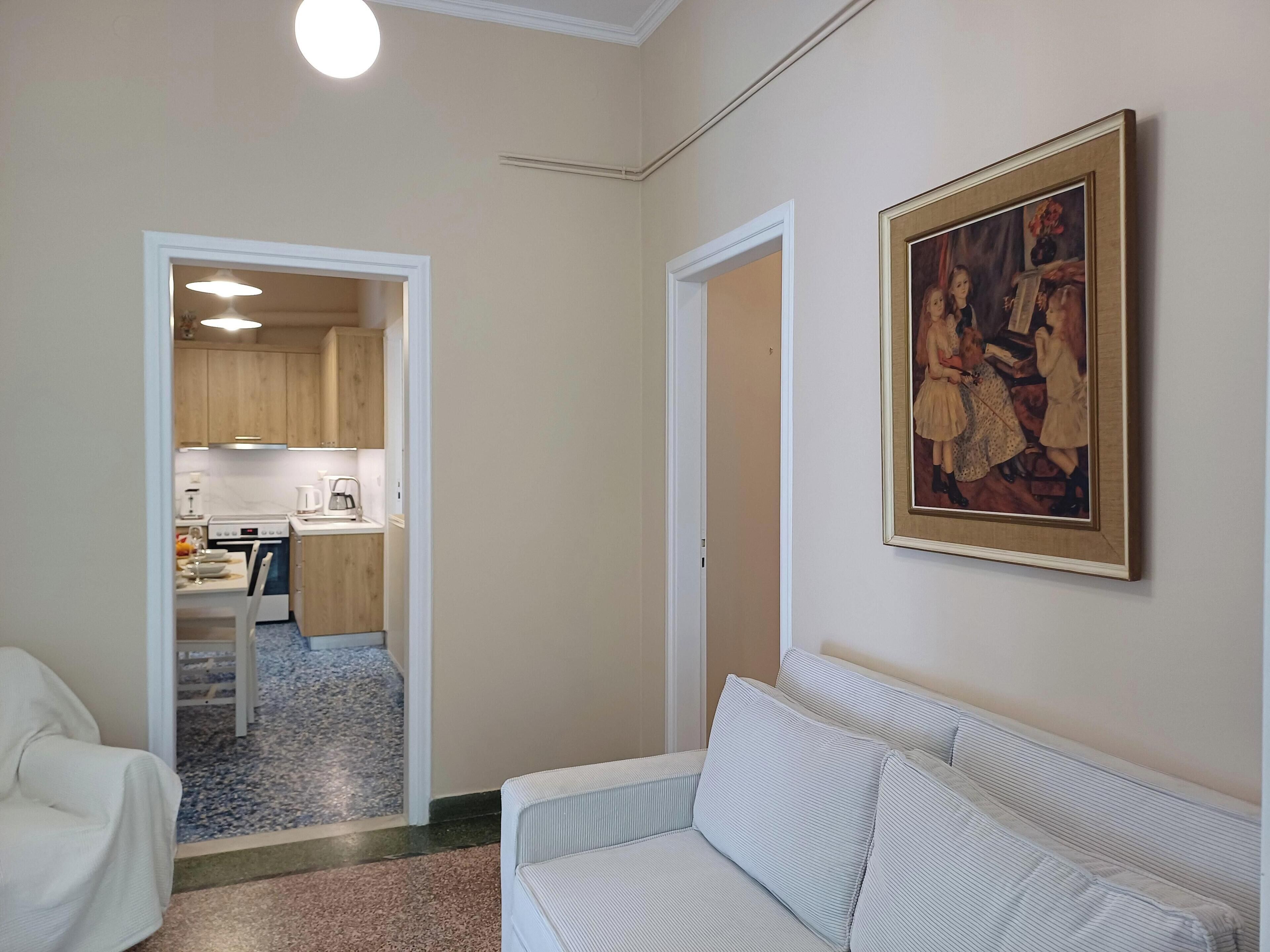 Un soggiorno con un divano, un quadro appeso al muro e una cucina visibile attraverso una porta aperta.