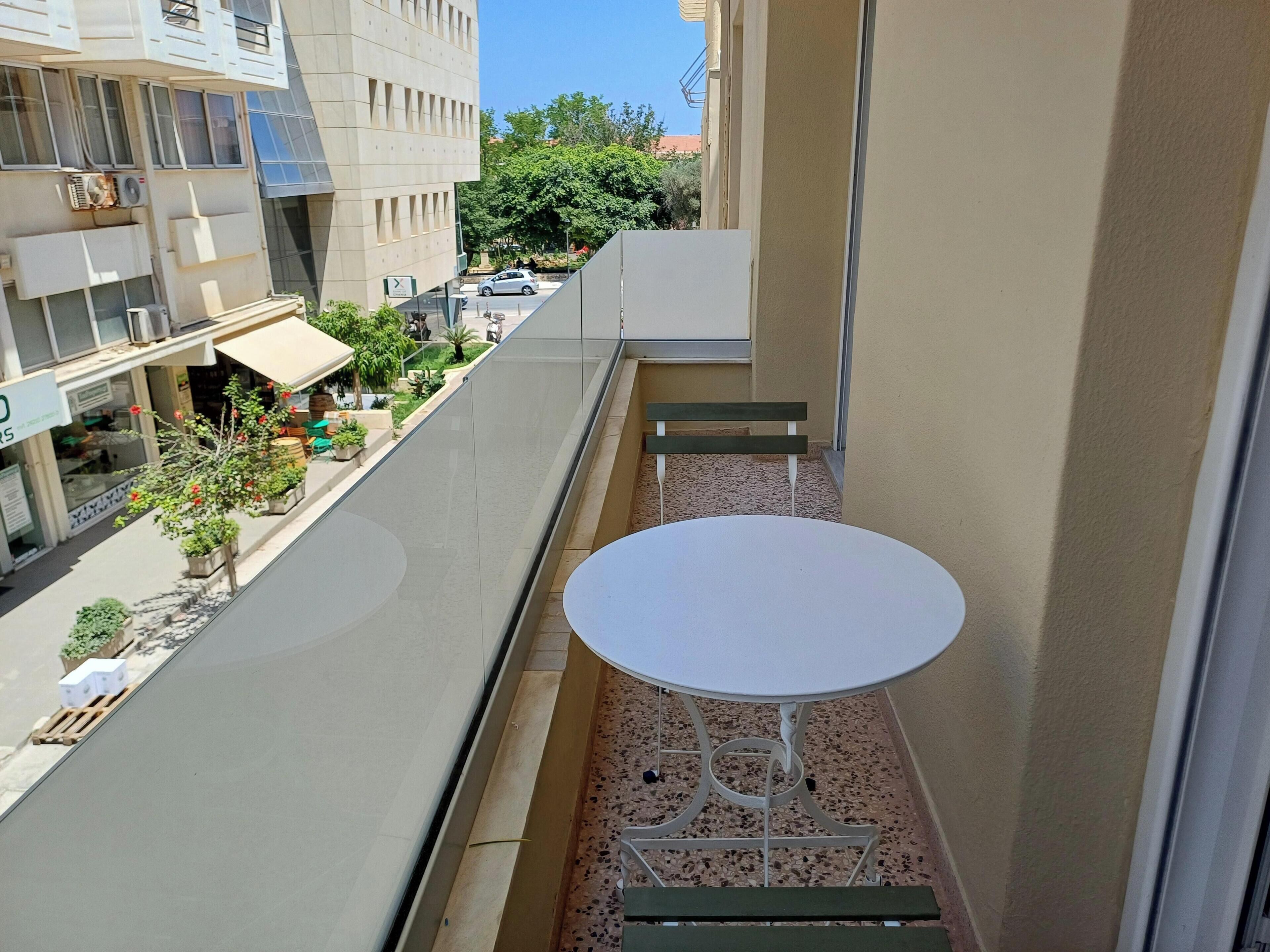A small balcony with a white table and two chairs, overlooking a street with shops and greenery.