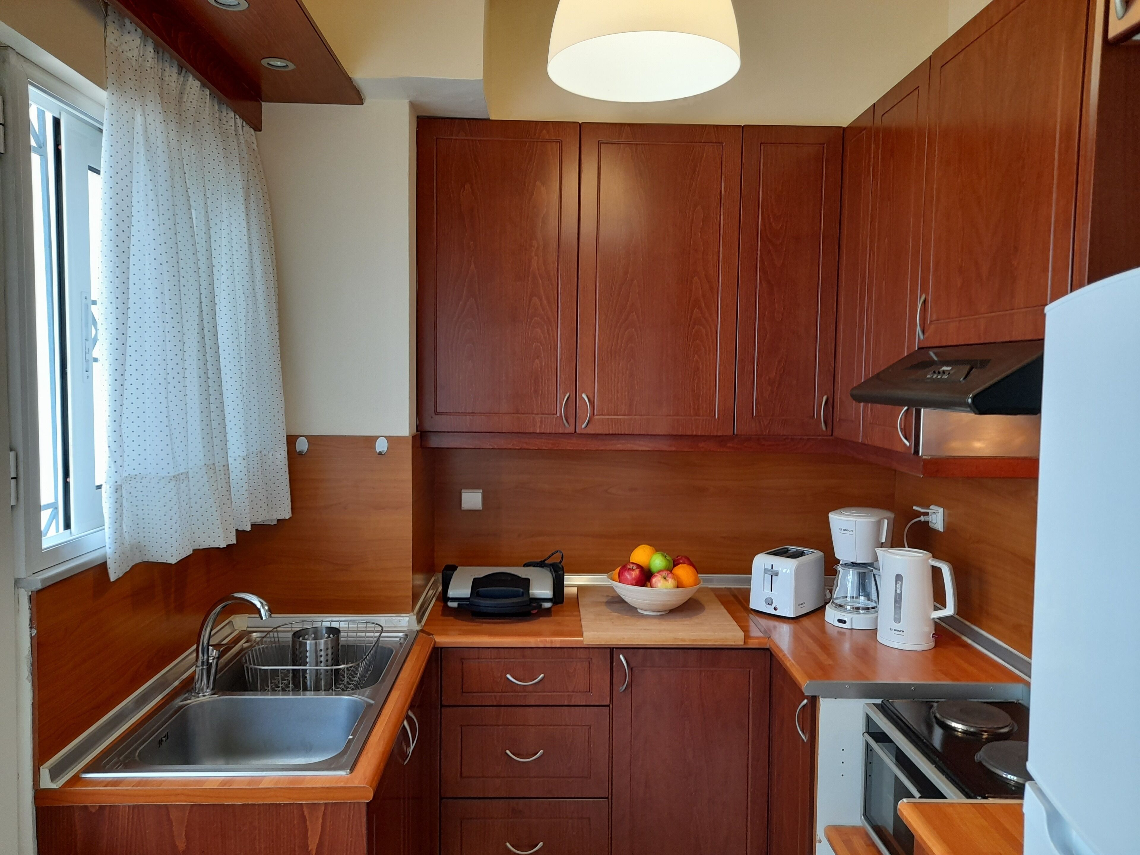 A compact kitchen with wooden cabinets, a stainless steel sink, a toaster, a coffee maker, and a bowl of fruit.