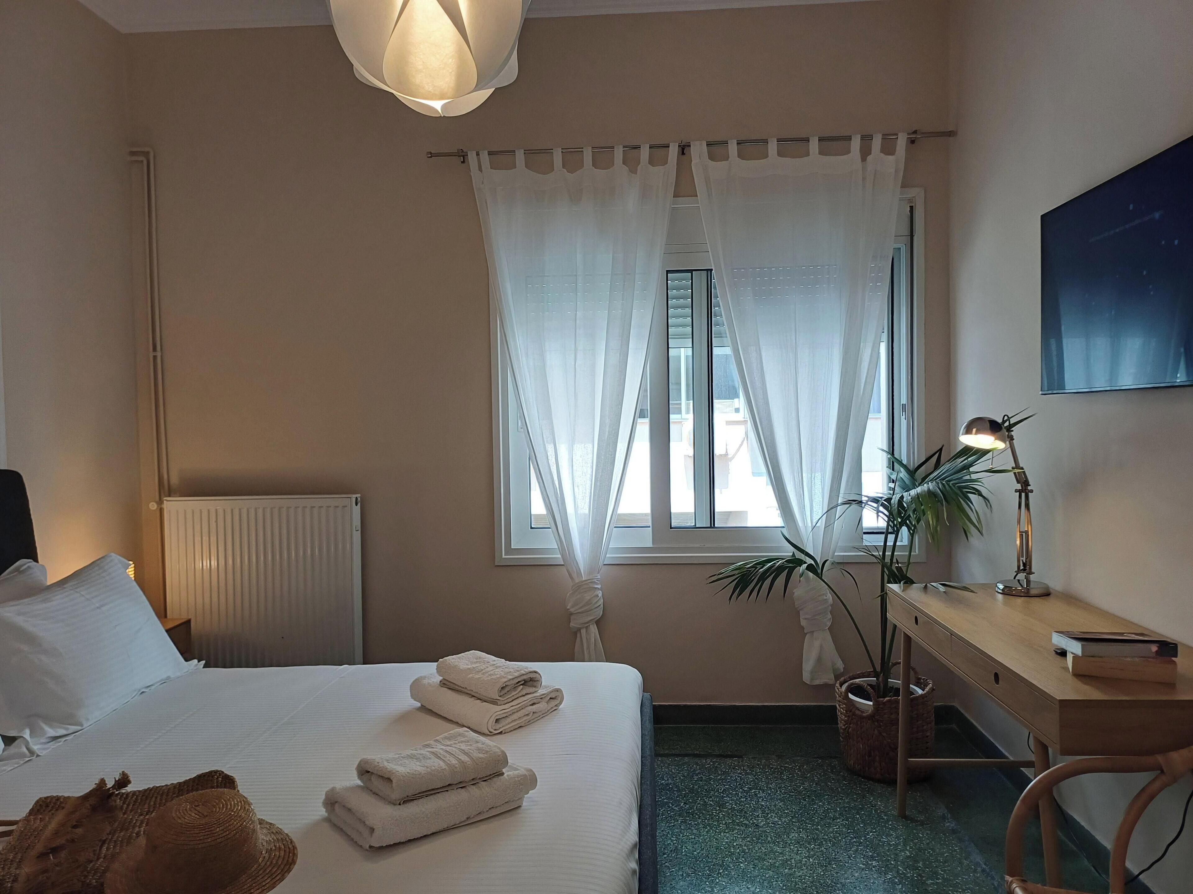 A hotel room with a bed, a desk, a lamp, a potted plant, and a window with sheer curtains.