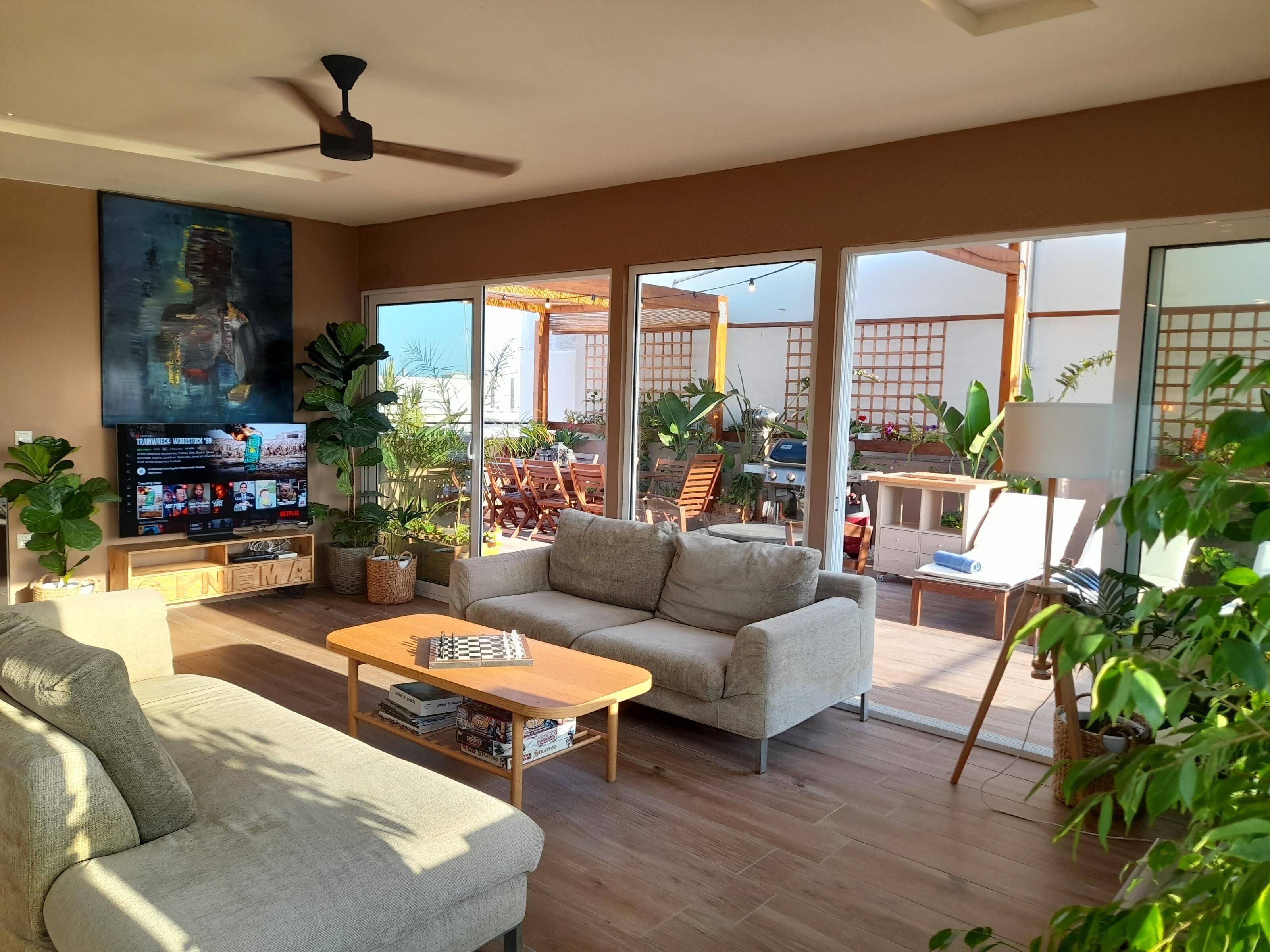 A spacious living room with a sofa, coffee table, and TV. There are potted plants and a balcony with outdoor furniture.