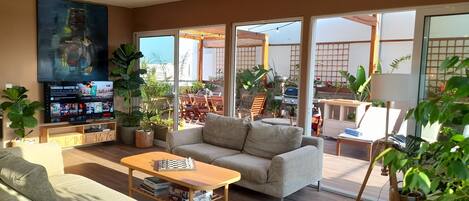 A spacious living room with a sofa, coffee table, and TV. There are potted plants and a balcony with outdoor furniture.