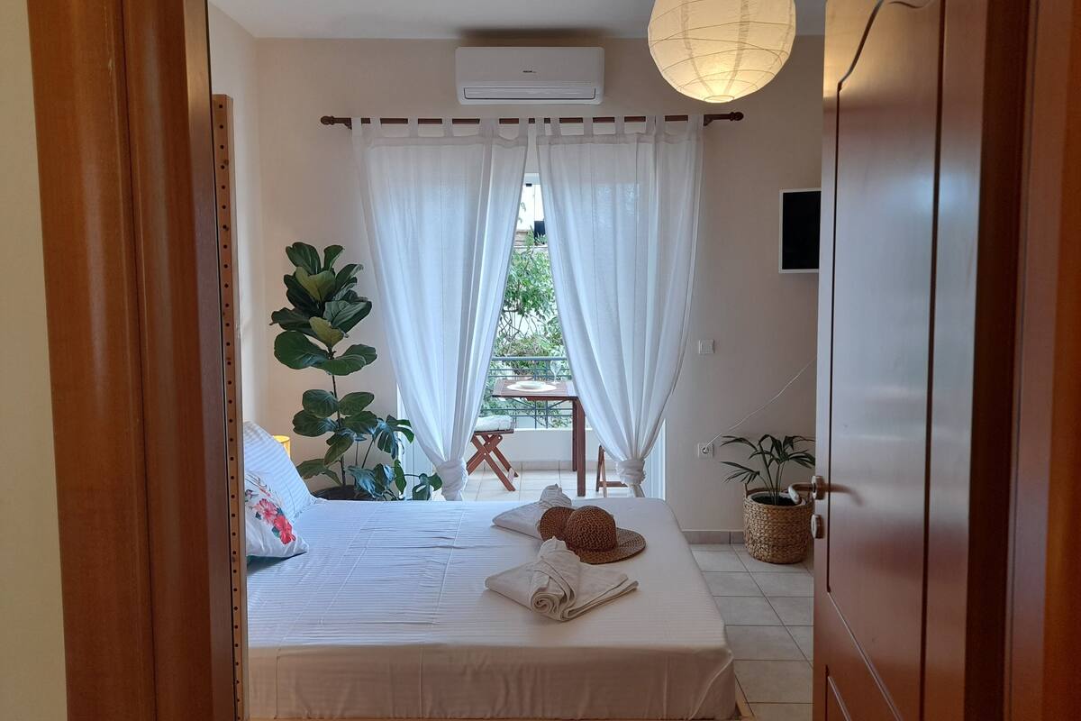 A bedroom with a bed, a bedside table, a potted plant, a window with curtains, and a view of the outdoors.