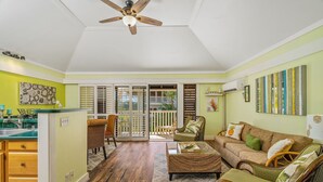 A spacious living area with a sofa, wicker chairs, and a dining table. A ceiling fan is visible, and the room has a large sliding glass door leading to a balcony. - Beautiful Poipu 1Br Condo with AC KP237 (Koloa)