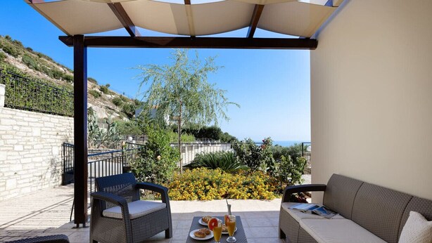Une terrasse extérieure avec des meubles en osier, une table garnie de nourriture et de boissons, et une vue sur la mer et les collines.