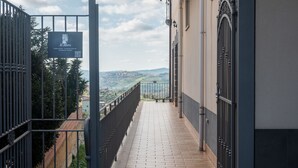 Un couloir au sol carrelé mène à une entrée fermée par un portail, d’où l’on peut admirer le paysage, et donne sur un bâtiment doté d’un balcon.