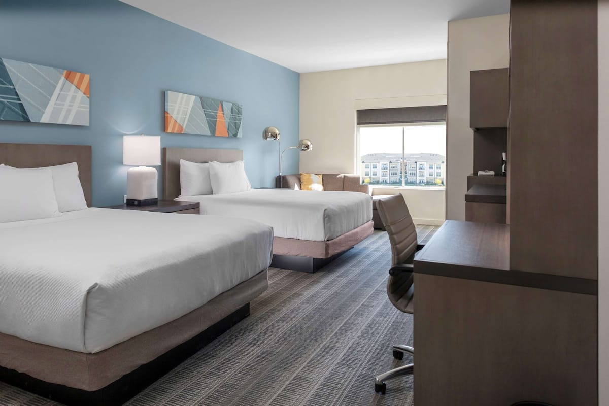 Ein Hotelzimmer mit zwei Betten, einem Schreibtisch, einem Sessel und einem großen Fenster mit Blick auf die Stadt.