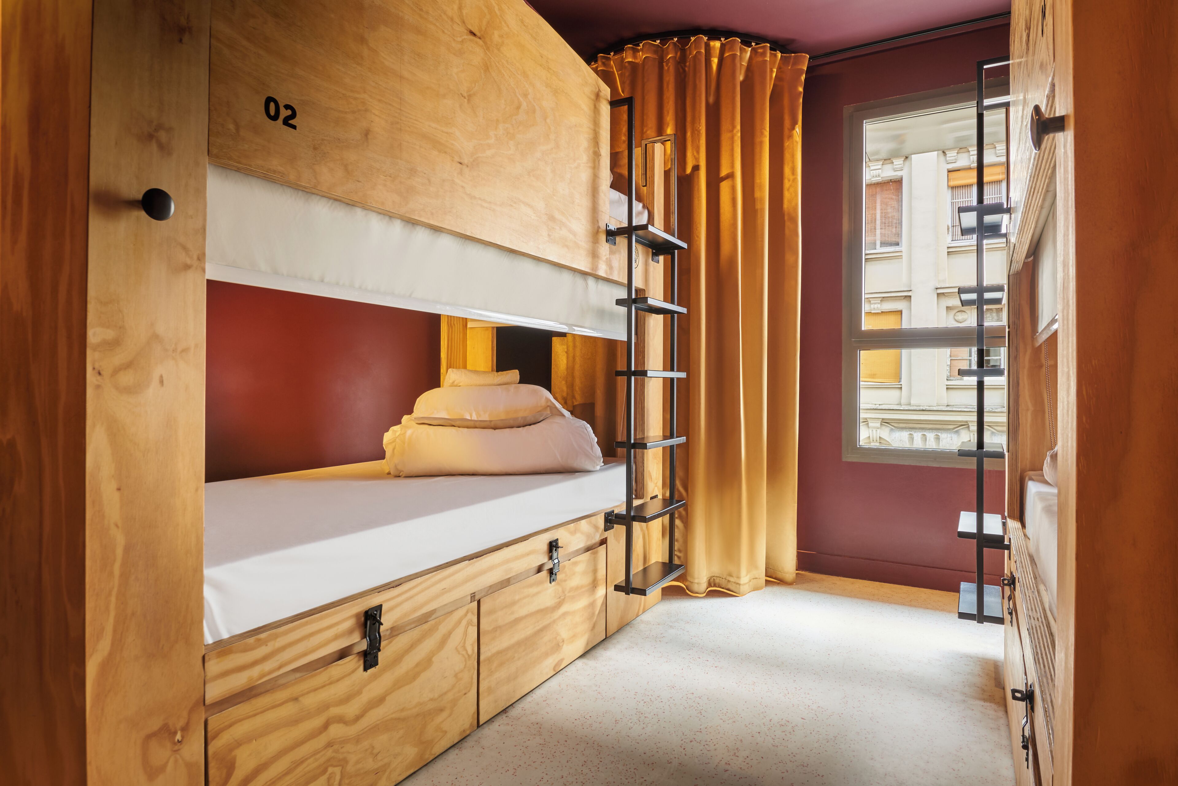 A bunk bed with a wooden headboard, a number '02' sign, a window with curtains, and a view of buildings outside.