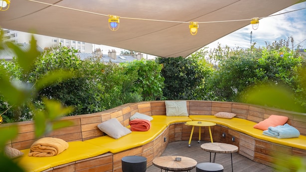 A covered outdoor seating area with wooden benches and tables, surrounded by greenery. - Jo&Joe Paris Nation - Hostel (Paris)
