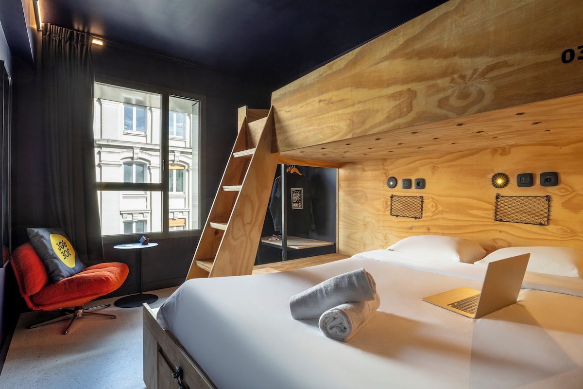 Moderni hotellihuone, jossa on sänky, kannettava tietokone, punainen tuoli, ikkuna verhoilla ja puinen seinä.