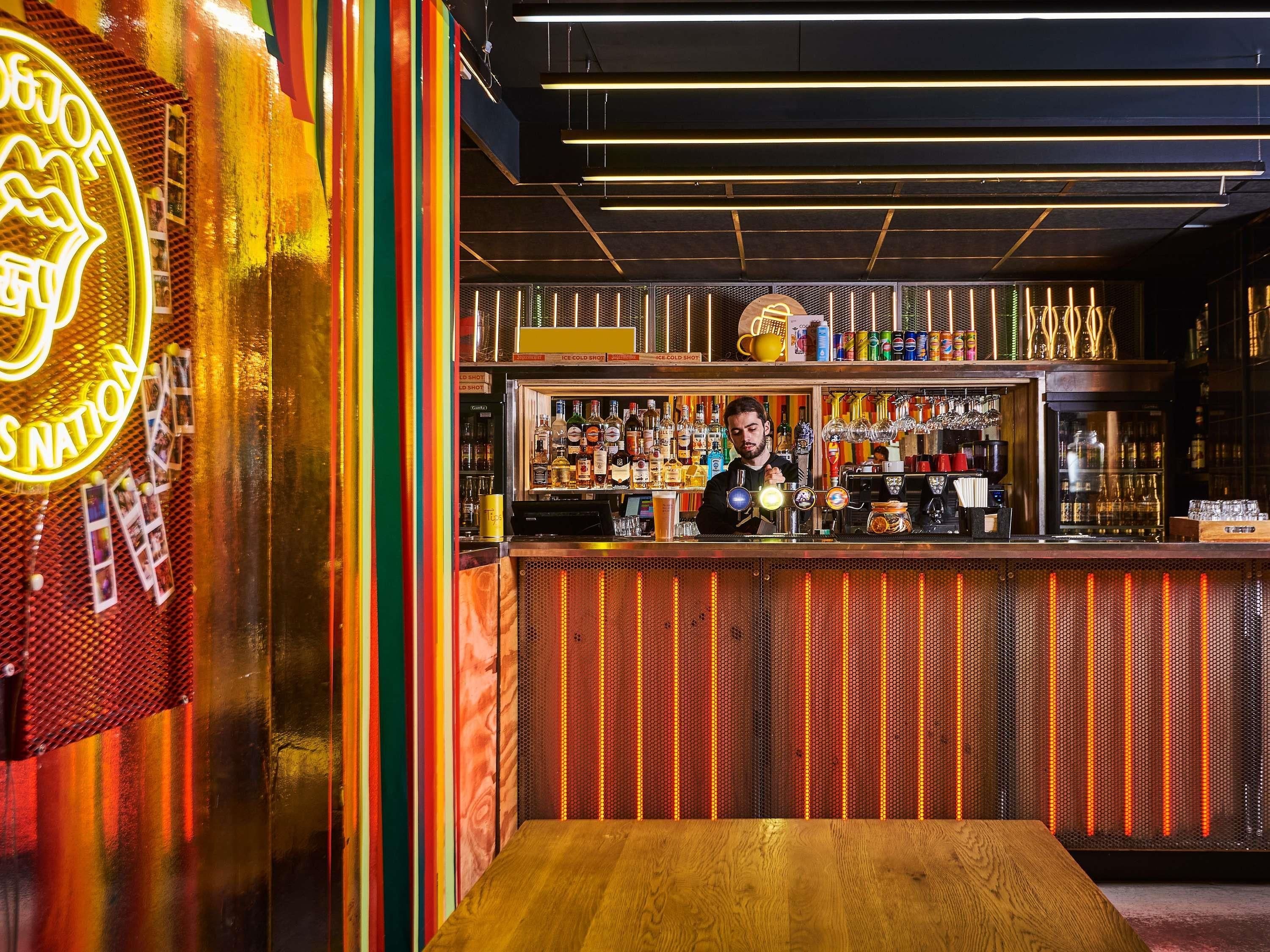 Eine Bar mit einer Neonreklame, einem Barkeeper und einer Auswahl an Flaschen und Gläsern auf Regalen.