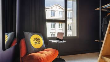 Une pièce moderne avec un seul fauteuil orange, une petite table ronde sur laquelle se trouve un ordinateur portable, une grande fenêtre donnant sur un immeuble et un porte-manteau.
