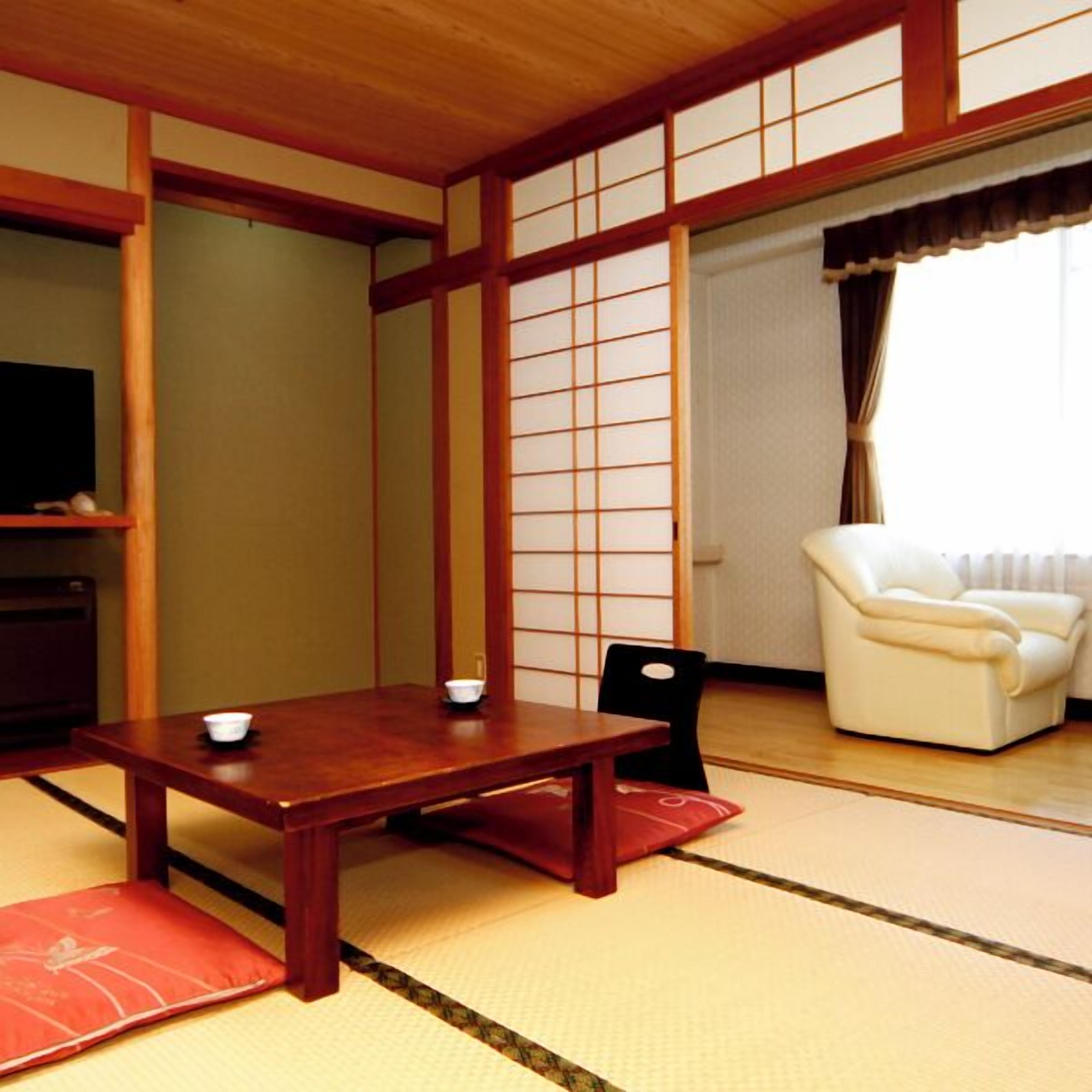 Una habitación japonesa tradicional con suelo de tatami, una mesa de madera baja, una silla negra, un sofá blanco y un televisor sobre una estantería.