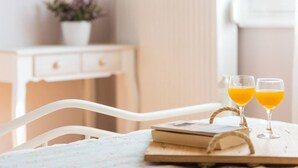 Ein weißer Stuhl, ein Holztisch mit einem Buch und zwei Gläsern Orangensaft, sowie eine weiße Kommode mit einer Pflanze im Hintergrund.