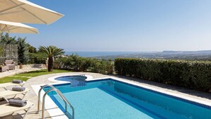 Ein Schwimmbecken mit Meerblick, umgeben von einer Hecke, und eine Liegefläche mit Stühlen und Sonnenschirm.