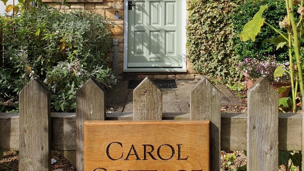 De ingang van een vakantiehuisje met een houten bord waarop 'CAROL COTTAGE' staat.