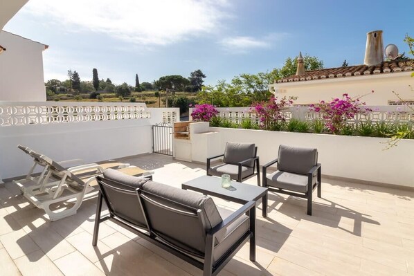 Une terrasse sur le toit, aménagée avec du mobilier d’extérieur, offrant une vue sur la campagne, et un bâtiment blanc doté d’une cheminée.