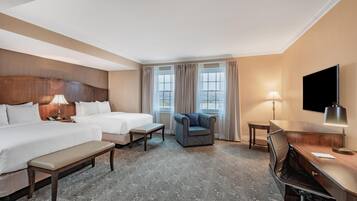 A hotel room with two beds, a desk, a chair, a bench, and a TV.