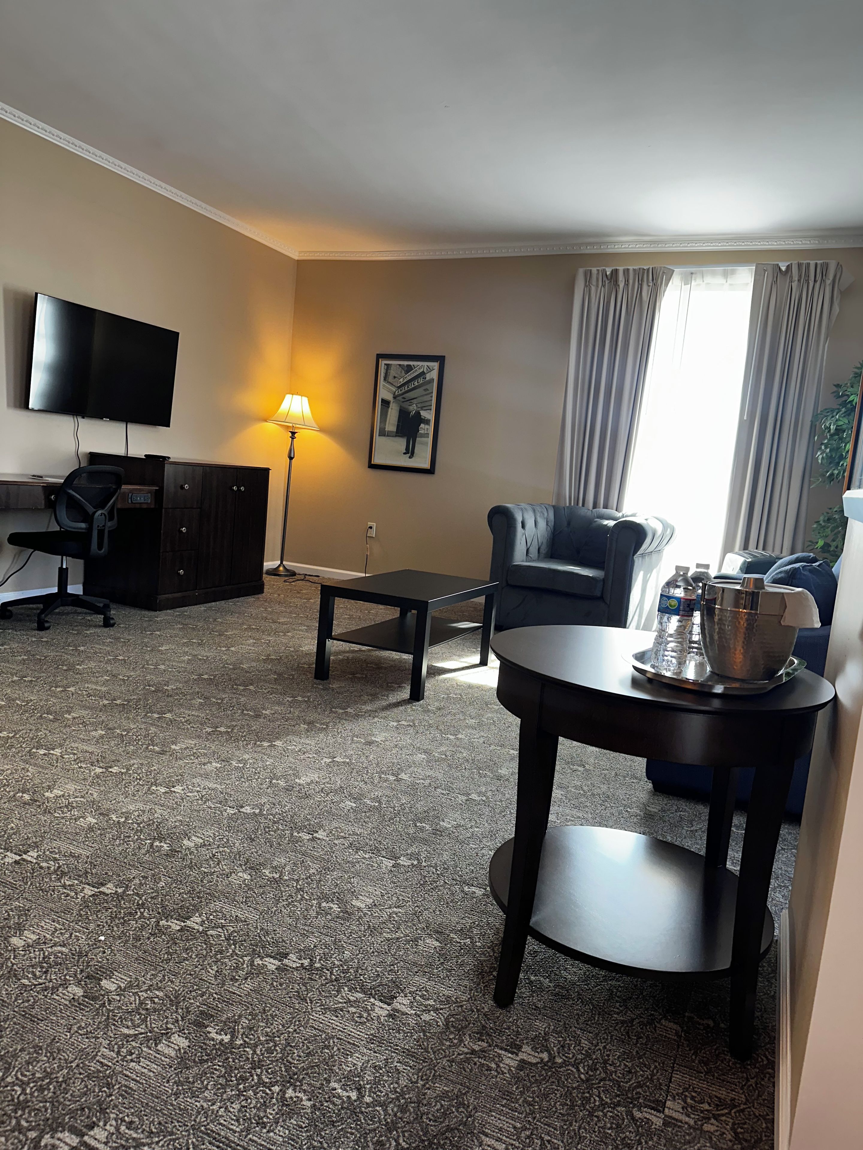 Habitación de hotel con televisor de pantalla plana, escritorio con silla, sofá, mesa de centro y mesa de comedor con jarra y botellas de agua.