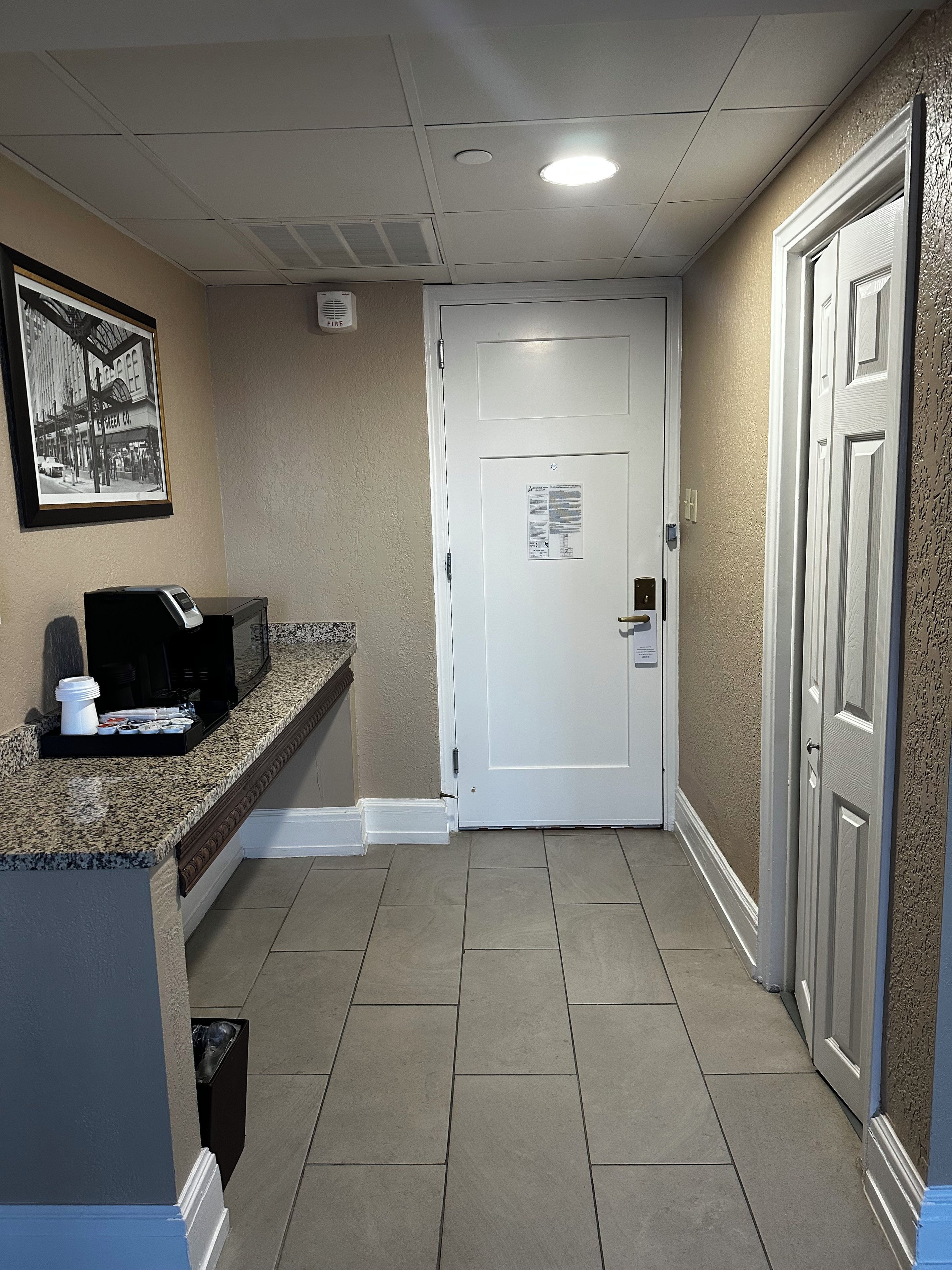 A hotel lobby with a reception desk, a framed picture on the wall, a white door with a fire exit sign, and a trash bin.