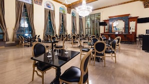 Un comedor amplio con techos altos, candelabros y ornamentada carpintería. Las mesas están dispuestas con sillas, y se observa una zona de bar al fondo.