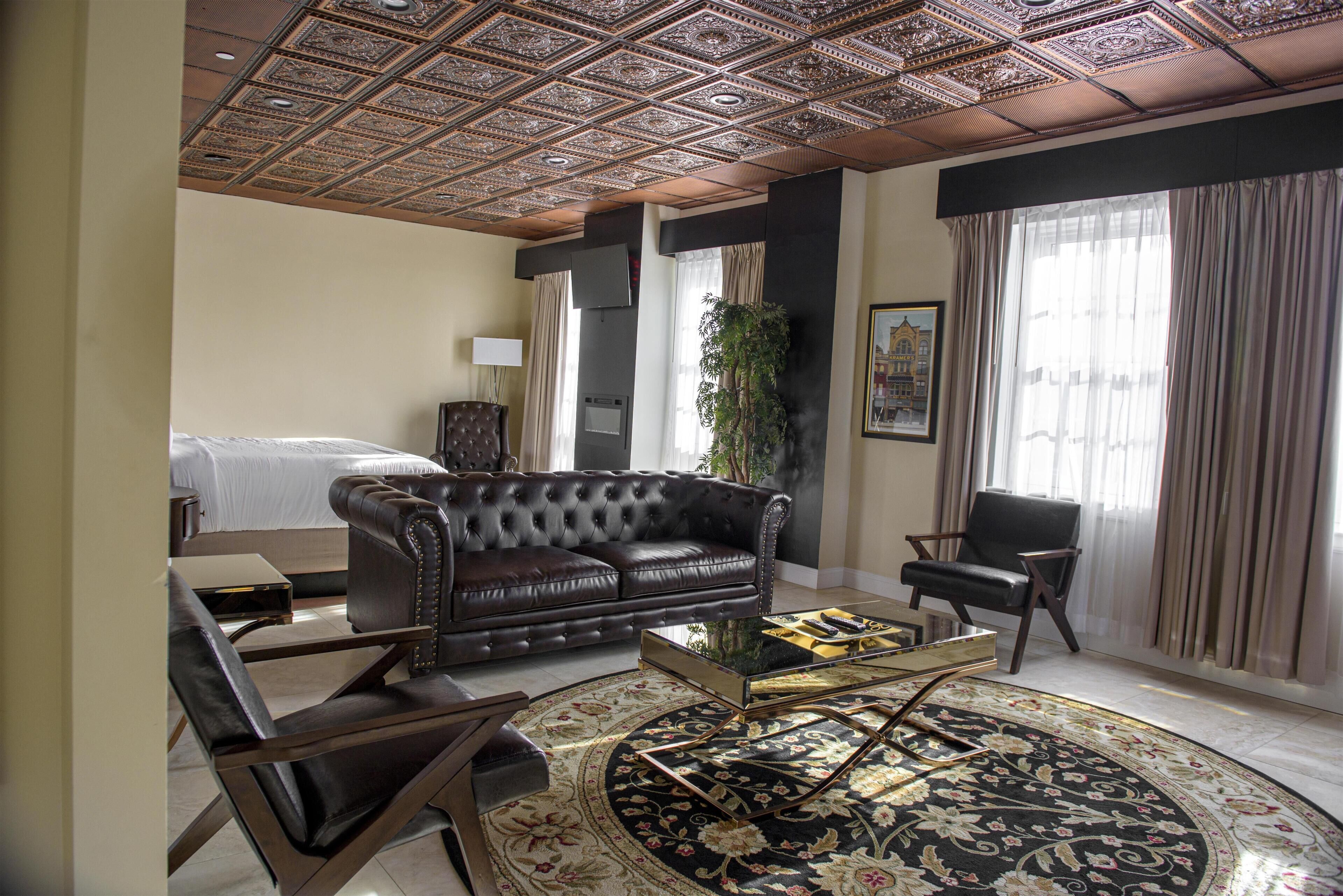 Habitación de hotel con sofá de cuero con capitonado, una mesa redonda, una cama, una silla, un ventanal con cortinas y un techo decorativo.